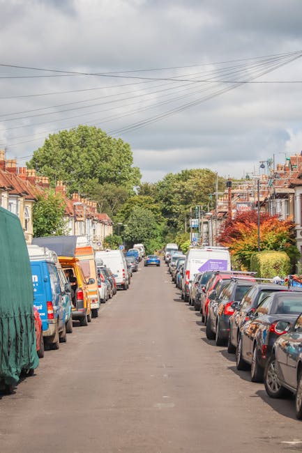 A residential street in Tooting during daytime showing parked cars lining both sides of the road alongside terraced houses with brick and stucco facades. Some houses have small front gardens with bushes and trees, including a large leafy green tree visible at the end of the street and smaller trees with red foliage closer to the foreground. Overhead, there are several power lines crossing the cloudy sky. The street appears narrow with limited space for passing vehicles, highlighting potential challenges for home relocation and furniture transport. The scene captures a typical urban environment where professional removals services, such as those provided by Movers Tooting, are often necessary to facilitate efficient packing and loading processes using moving equipment like trolleys and vans, to avoid delays and extra costs associated with parking restrictions or street congestion.
