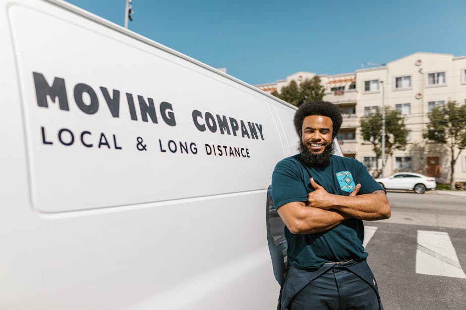 A man wearing a white polo shirt, jeans, and a black cap stands inside the back of a large white moving van, holding a cardboard box with both hands. The interior of the van is filled with neatly stac